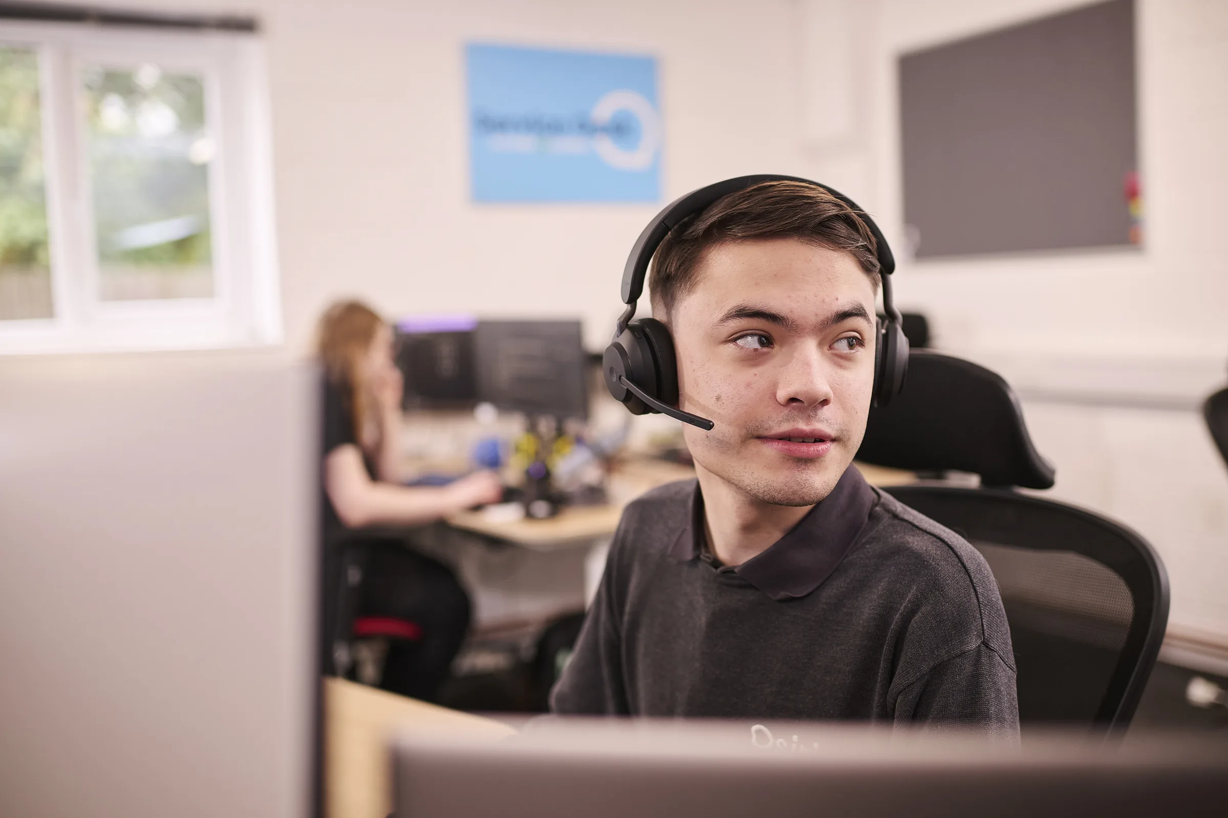 An Osiris IT engineer wearing a headset at his desk in the Norwich office, with a colleague working behind him.